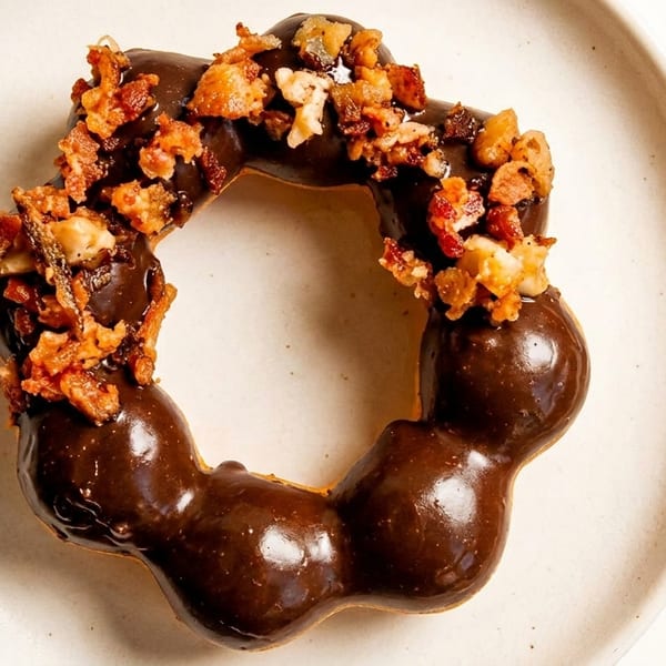 A chocolate-glazed mochi donut topped with crispy bacon bits and nuts on a white plate.