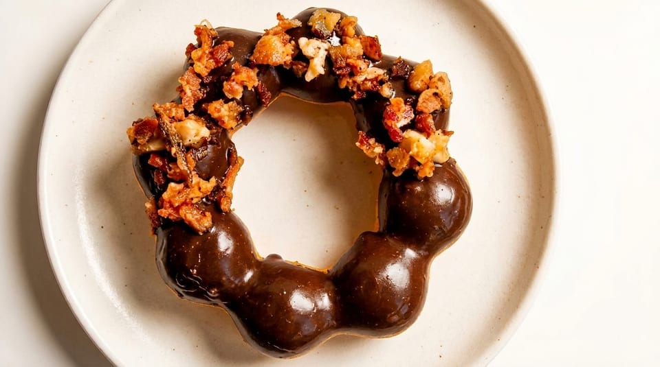 A chocolate-glazed mochi donut topped with crispy bacon bits and nuts on a white plate.