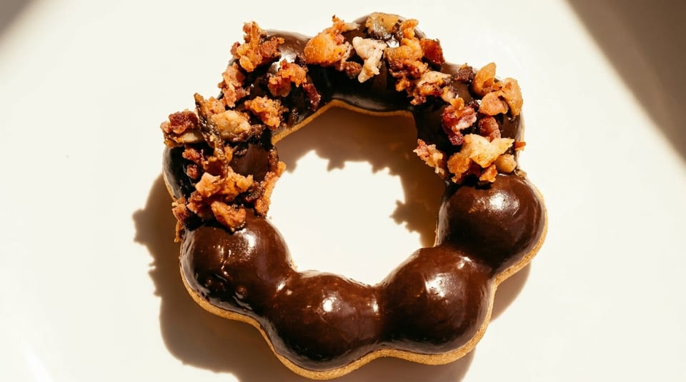 A chocolate-glazed mochi donut topped with crispy bacon bits, casting a shadow on a white surface.