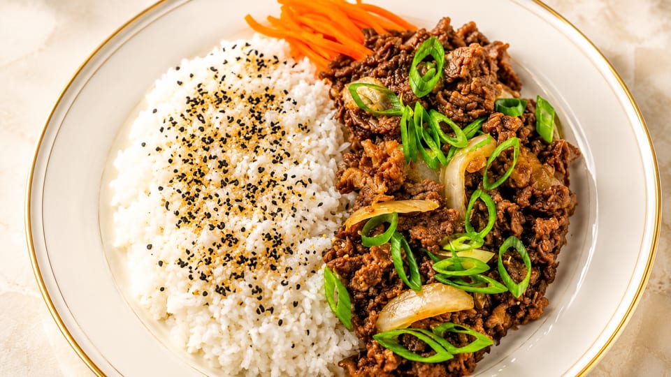 Bulgogi beef with white rice topped with sesame seeds and shredded carrots on a white plate.